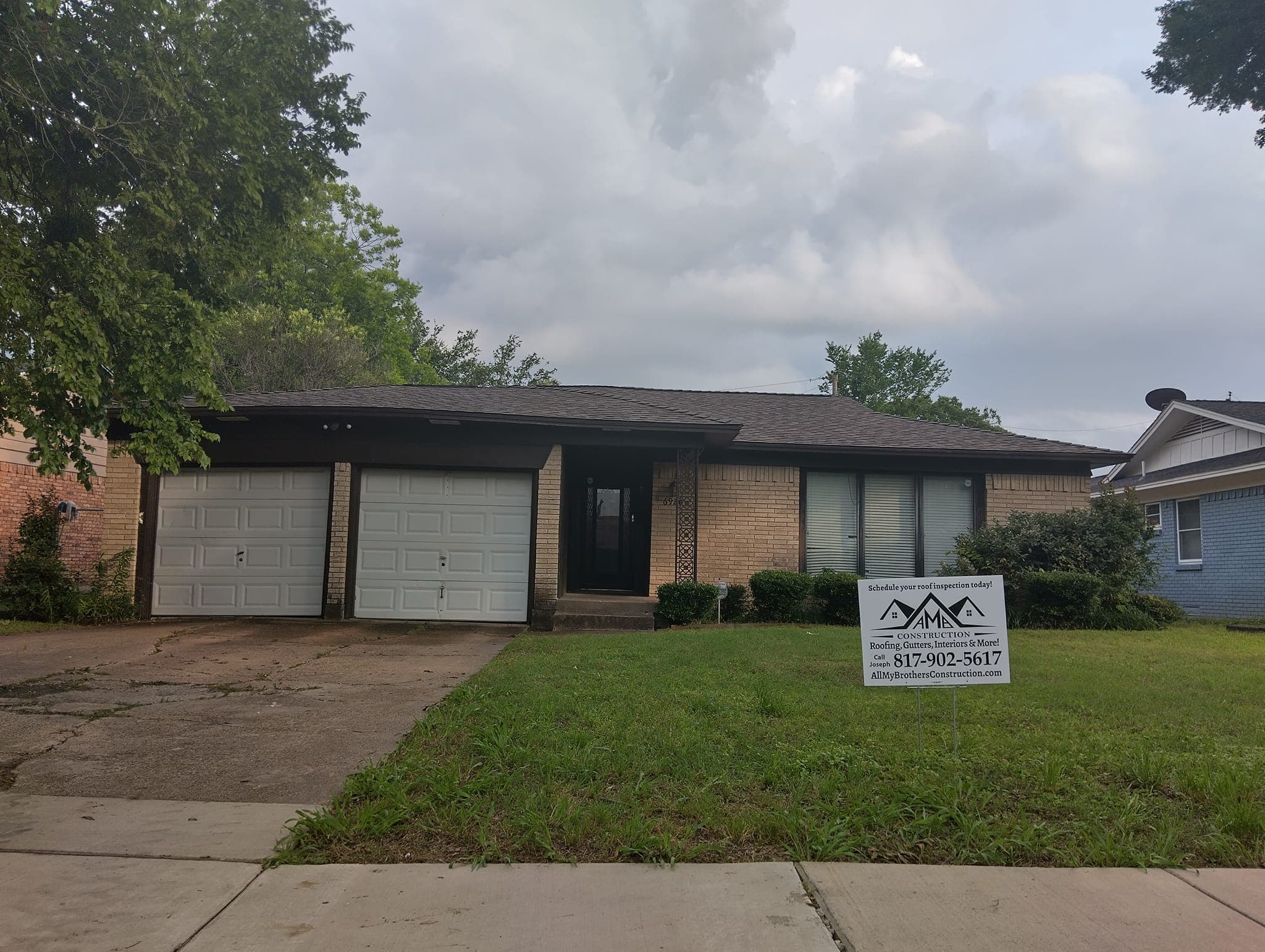 Single-story brick home with a for sale sign, two-car garage, and green lawn under cloudy sky.